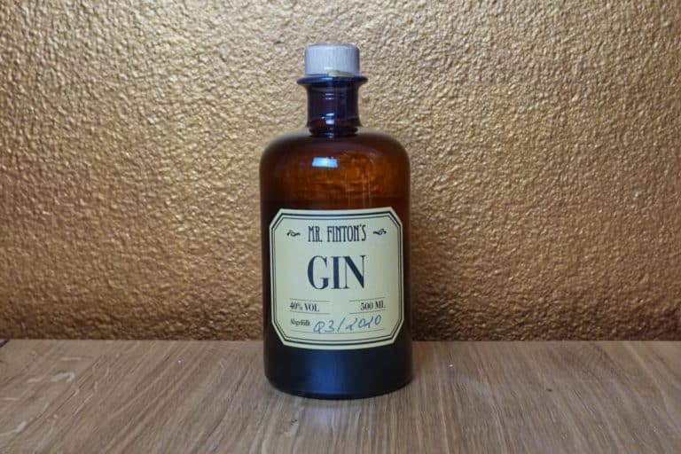 Braune Glasflasche mit der Aufschrift „Mr. Fintons Gin“, 500 ml, 40 % vol. Die Flasche hat einen Korkverschluss und steht auf einer Holzunterlage mit goldfarbenem, strukturiertem Hintergrund. Diese edle Präsentation von „Mr. Fintons Gin“ unterstreicht die Qualität, die Sie bei Lidl finden.