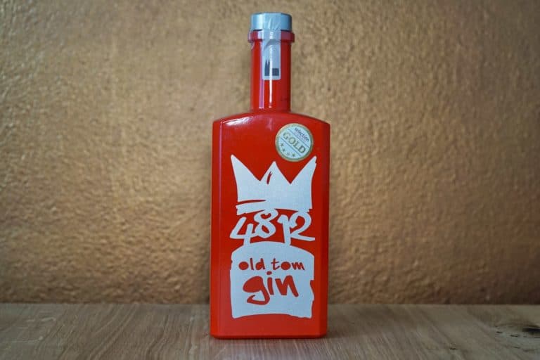 Eine rote Flasche „48K Tom Gin“ mit einem weißen Kronenmotiv und einem goldenen Auszeichnungsaufkleber steht auf einer Holzfläche vor einer hellbraunen, strukturierten Wand.