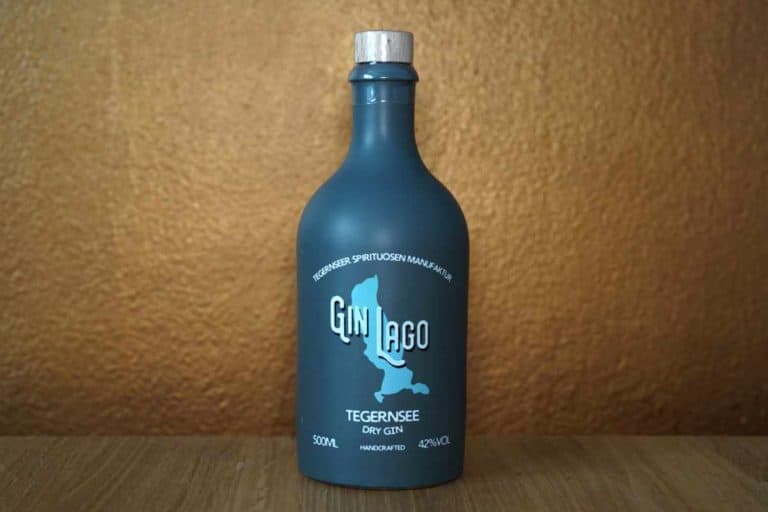 Eine blaue Flasche „Gin Lago Tegernsee Dry Gin“ steht auf einer Holzunterlage vor einem strukturierten braunen Hintergrund. Das Etikett zeigt weißen Text, die Silhouette eines Sees und präsentiert stolz das unverwechselbare „Gin Lago“-Logo.