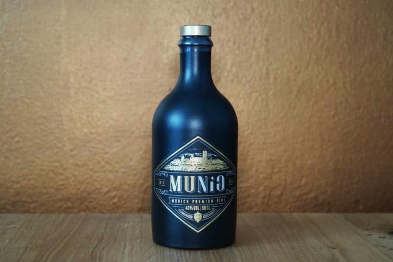 Eine dunkelblaue Flasche „Munich Gin“ mit der Aufschrift „Munio“ in gold-weißer Schrift steht auf einer Holzunterlage vor einem hellbraunen Hintergrund.