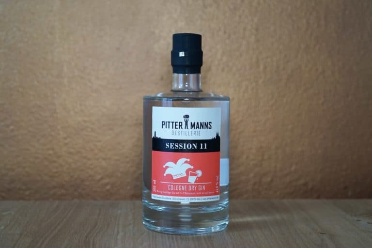 Eine Flasche „Pittermanns Distillery Session 11 London Dry Gin“ steht auf einer Holzfläche vor einer hellbraunen Wand. Das Etikett ist weiß und orange mit schwarzer Schrift und Grafiken und bringt diesen außergewöhnlichen London Dry Gin perfekt zur Geltung.