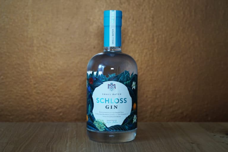 Eine Flasche Schloss Gin mit blauem Verschluss und illustriertem Etikett steht auf einer Holzfläche vor einer braunen, strukturierten Wand und verkörpert die raffinierte Eleganz, die Schloss Gin auszeichnet.
