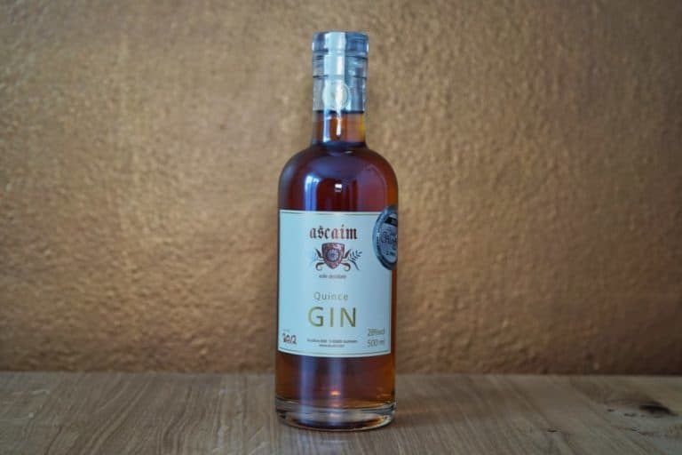 Eine Flasche Ascaim Quince Gin steht auf einer Holzfläche vor einer strukturierten braunen Wand; das Etikett ist deutlich zu erkennen, und das charakteristische Siegel von Ascaim Quince Gin ist unversehrt.