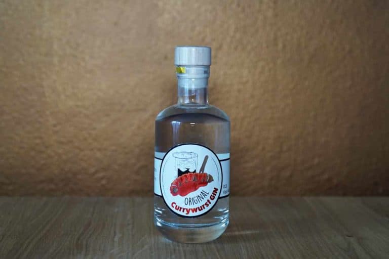 Auf einer Holzfläche steht eine durchsichtige Glasflasche mit einem silbernen Verschluss. Auf dem Etikett steht „Original Currywurst Geist“ und es zeigt eine Abbildung einer Currywurst mit Pommes frites und einem Zahnstocher, was an den Charme klassischer deutscher Straßenküche erinnert. Dieses einzigartige Getränk, das an innovative Kreationen wie den Currywurst-Gin erinnert, wird vor dem Hintergrund einer schlichten braunen Wand präsentiert.