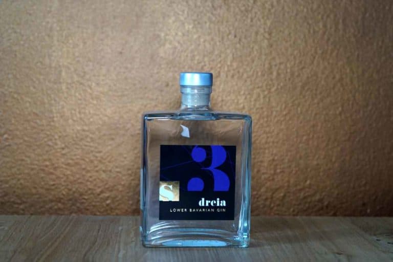 Eine quadratische Glasflasche „Dreia Lower Bavarian Gin“ mit blauem Verschluss, die auf einer Holzfläche vor einer braunen, strukturierten Wand steht.
