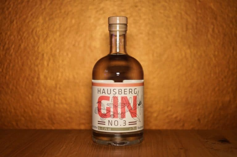 Eine durchsichtige Flasche Hausberg Gin Nr. 3 steht auf einer Holzunterlage vor einem goldfarbenen, strukturierten Hintergrund. Auf dem Etikett ist der markante Name „Hausberg Gin Nr. 3“ in roter und schwarzer Schrift zu sehen, ergänzt durch elegante Illustrationen von Pflanzen.