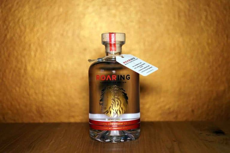 Eine klare Glasflasche „Löwensenf ROARING Gin“ mit rotem Verschluss und Etikett, auf dem ein Löwenmotiv abgebildet ist, steht auf einer Holzunterlage vor einem strukturierten goldenen Hintergrund. Am Flaschenhals hängt ein kleines Etikett.