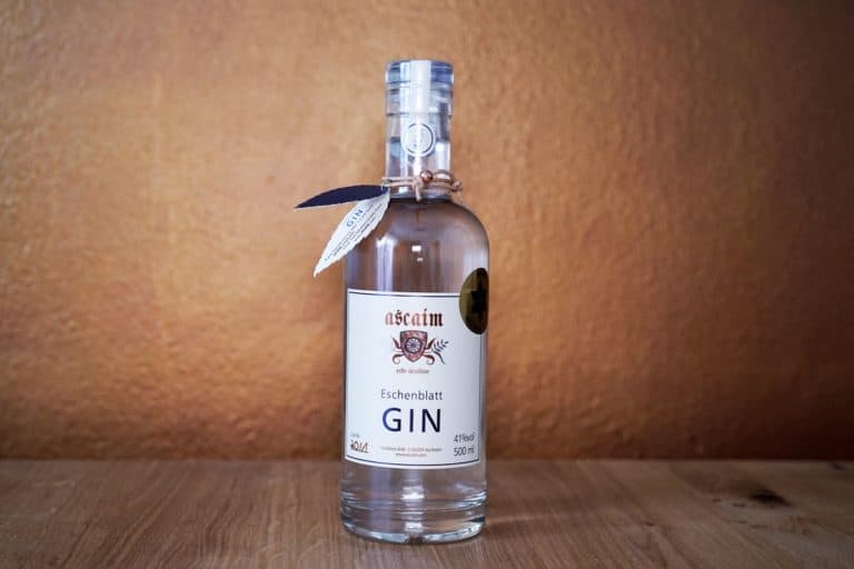 Eine klare Glasflasche „Ascaim Eschenblatt Gin“ mit weißem Etikett steht auf einer Holzfläche vor einer strukturierten braunen Wand.
