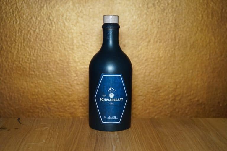 Eine dunkelblaue Flasche mit der Aufschrift „Schwarzbart Gin“ und einem Korkverschluss steht auf einer Holzunterlage vor einem strukturierten goldbraunen Hintergrund und strahlt eine geheimnisvolle Eleganz aus, die dem vornehmen Charakter des Schwarzbart Gin gerecht wird.