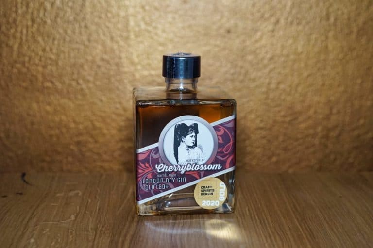 Eine quadratische Glasflasche „Cherryblossom London Dry Gin“ mit schwarzem Verschluss steht auf einer Holzfläche vor einem strukturierten goldenen Hintergrund, der an einen eleganten Gin-Reifekeller erinnert. Auf dem Etikett sind das Porträt einer Frau und ein goldenes Abzeichen mit der Aufschrift „Craft Spirits Berlin 2020“ zu sehen.