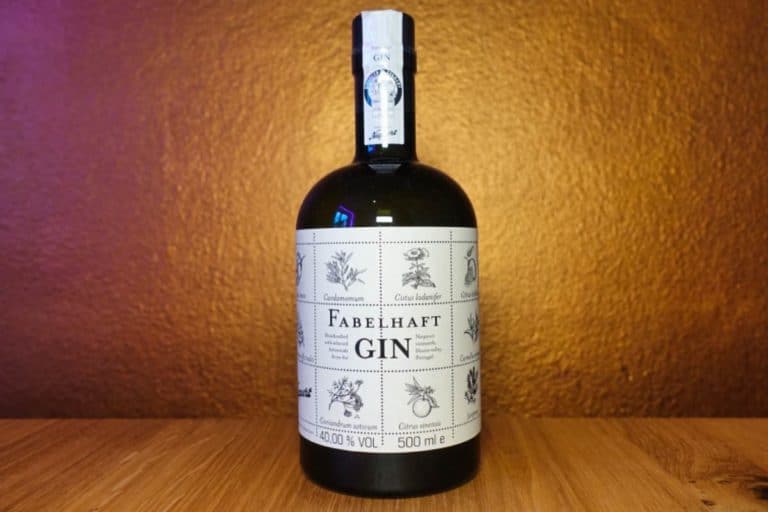 Eine Flasche „Fabelhaft Gin“ mit einem weißen Etikett, das botanische Illustrationen zeigt, steht auf einer Holzunterlage vor einem warmen, bräunlich-goldenen Hintergrund und bringt den handwerklichen Charakter von „Fabelhaft Gin“ perfekt zum Ausdruck.