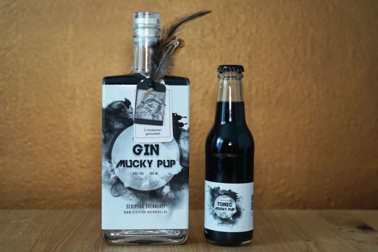 Auf einer Holzfläche vor einem goldenen Hintergrund steht eine Flasche „Mucky Pup Gin“, an deren Flaschenhals eine Feder befestigt ist, neben einer kleinen Flasche mit der Aufschrift „Tonic Mucky Pup“.