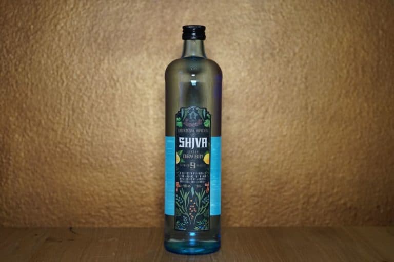 Eine Flasche Shiva London Dry Gin (Lidl) steht auf einer Holzunterlage vor einem strukturierten, goldbraunen Hintergrund. Das Etikett zeigt farbenfrohe botanische Illustrationen und oben ein grünes Logo.