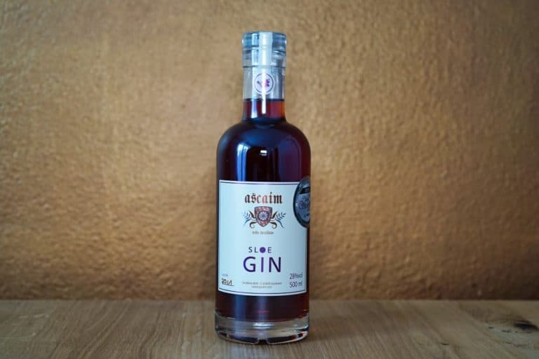 Eine Flasche Ascain Sloe Gin mit cremefarbenem Etikett steht auf einer Holzunterlage vor einem strukturierten, hellbraunen Hintergrund und unterstreicht die handwerkliche Qualität, für die Ascain Gin bekannt ist.