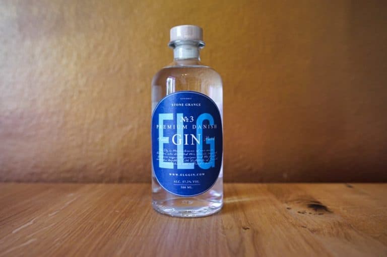 Eine klare Glasflasche mit „Elgin Premium Danish Gin“, auch bekannt als „ELG Gin No. 3“, mit einem blauen Etikett steht auf einer Holzunterlage vor einem braunen Hintergrund.