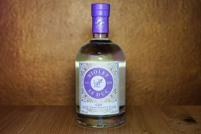 Eine durchsichtige Flasche „Violet Le Duc Gin“ mit einem verzierten, violett-weißen Etikett und Verschluss, die auf einer Holzunterlage vor einem braunen Hintergrund steht.