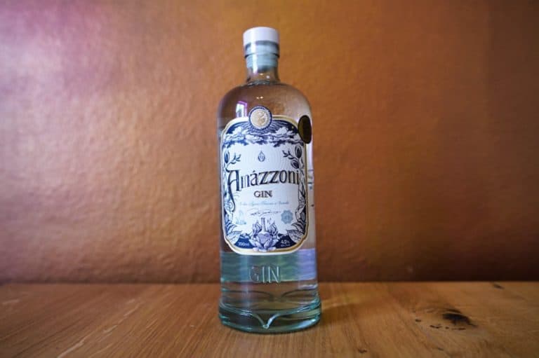 Eine klare Glasflasche Amazzoni Gin mit einem dekorativen weißen Etikett steht auf einer Holzunterlage vor einem braunen Hintergrund und unterstreicht die Eleganz und handwerkliche Qualität von Amazzoni Gin.
