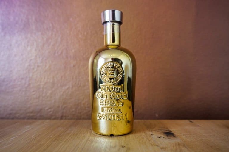 Eine glänzende, goldene Flasche „Gold Gin“ mit der Prägung „700 ml Gin Gold 999,9“ steht aufrecht auf einer Holzunterlage vor einem braunen Hintergrund.