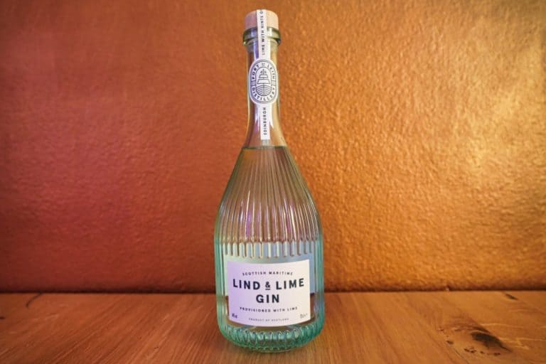 Eine klare, geriffelte Glasflasche „Lind and Lime Gin“ mit einem hellblau-weißen Etikett steht auf einer Holzfläche vor einer strukturierten, bronzefarbenen Wand.