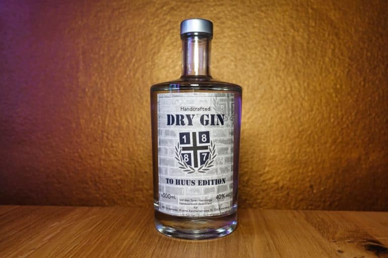 Eine Flasche „1887 Handcrafted Dry Gin – To Huus Edition“ steht elegant auf einer Holzunterlage vor einer goldbraunen, strukturierten Wand; ihr handgefertigtes Etikett spiegelt die Hingabe wider, die in jede einzelne Kleinserie fließt.
