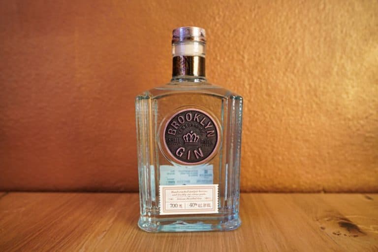 Eine durchsichtige Glasflasche Brooklyn Gin mit silbernem Verschluss steht auf einer Holzunterlage vor einer goldbraunen Wand. Auf dem markanten Etikett sind der Markenname „Brooklyn Gin“ und der Alkoholgehalt angegeben.