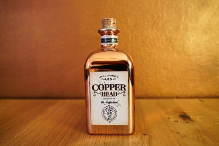 Eine glänzende, kupferfarbene Flasche Copperhead Gin mit Korkverschluss steht auf einer Holzunterlage vor einem passenden kupferfarbenen Hintergrund und unterstreicht so die unverwechselbare Eleganz des Copperhead Gin.