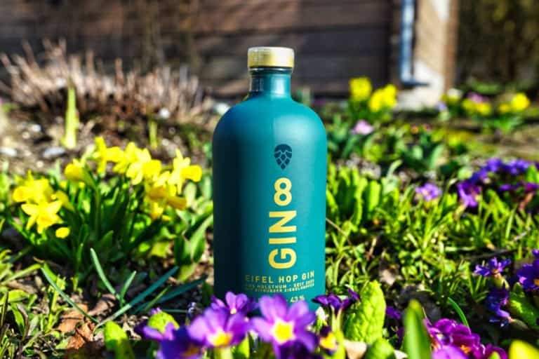 Eine blaugrüne Flasche „Gin 8 Eifel Hop Gin“ steht in einem Garten inmitten blühender gelber und violetter Blumen, im Hintergrund ist ein verschwommenes Holzgebäude zu sehen.