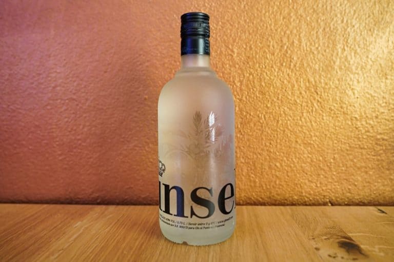 Eine Milchglasflasche „Ginself Gin“ mit schwarzem Verschluss steht auf einer Holzfläche vor einem warmen, strukturierten orangefarbenen Hintergrund. Das Etikett der Flasche ist teilweise zu sehen und zeigt die Buchstaben „nse“.
