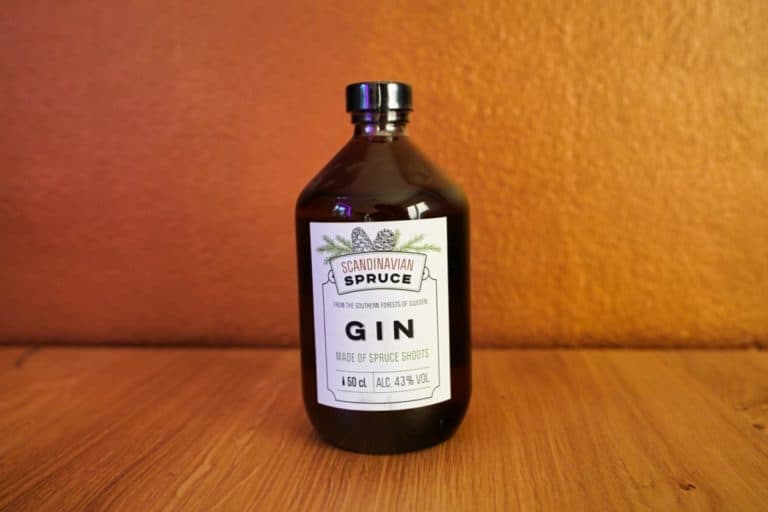 Auf einer Holzfläche vor orangefarbenem Hintergrund steht eine braune Flasche „Scandinavian Spruce Gin“. Auf dem Etikett ist vermerkt, dass dieser einzigartige Gin aus Fichtentrieben hergestellt wird, 500 ml enthält und einen Alkoholgehalt von 43 % aufweist.
