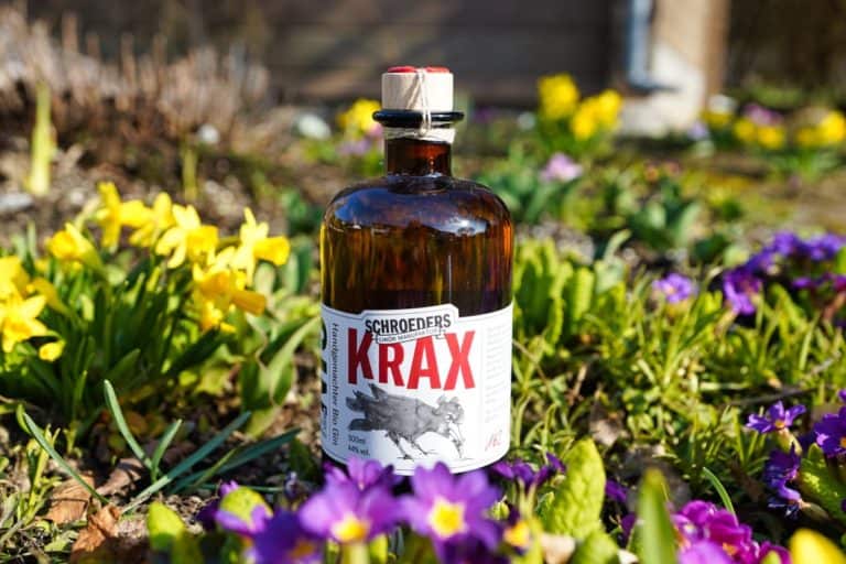 Eine braune Glasflasche mit der Aufschrift „Schroeders Krax Gin“ steht inmitten blühender gelber und violetter Blumen auf einer grasbewachsenen Freifläche.