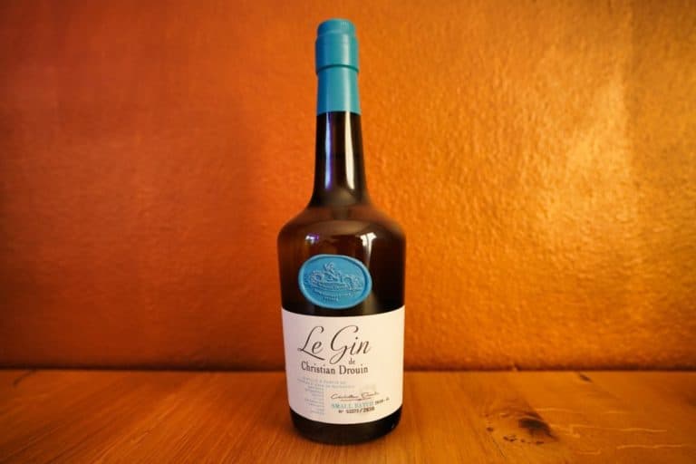Eine Flasche „Le Gin de Christian Drouin“ mit blauem Verschluss und Etikett steht auf einer Holzunterlage vor einem orangefarbenen Hintergrund.