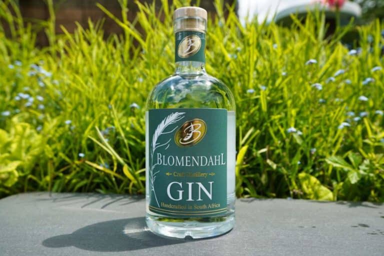 Eine Flasche Blomendahl Gin steht im Freien auf einer grauen Fläche, im unscharfen Hintergrund sind grünes Gras und kleine blaue Blumen zu sehen, was die natürliche Eleganz des Blomendahl Gin in dieser ruhigen Umgebung unterstreicht.