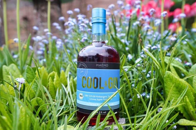 Eine Flasche Maëmo Cool & Gin (ma mo Cool Gin) steht inmitten von saftig grünem Gras und kleinen blauen Blüten, im Hintergrund sind verschwommene rote Blüten zu sehen.