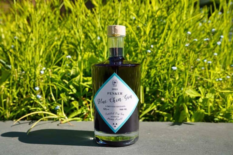 Eine Flasche „Penker Blue Chin Gin“ mit einem rautenförmigen Etikett steht im Freien auf einer grauen Fläche, im Hintergrund sind saftig grünes Gras und kleine blaue Blumen zu sehen.