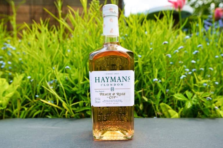 Eine Flasche Gin „Haymans London Peach & Rose Cup“ steht im Freien auf einer grauen Fläche, im Hintergrund sind grünes Gras und kleine blaue Blumen verschwommen zu sehen.