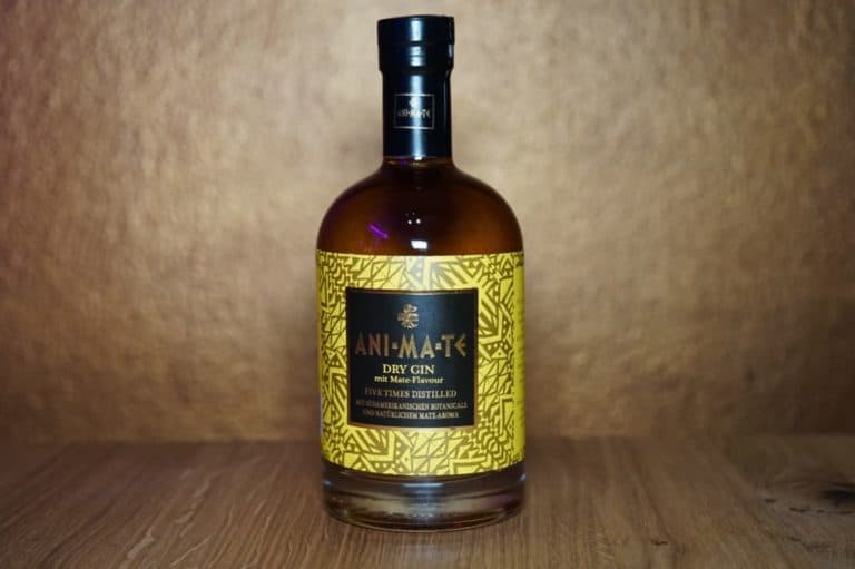 Eine Flasche „Ani Ma Te Gin“ mit einem gelb gemusterten Etikett steht auf einer Holzfläche vor einem braunen Hintergrund.