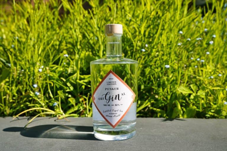 Auf einer Steinfläche steht eine durchsichtige Glasflasche mit Gin der Marke Penker, im Hintergrund sind saftiges grünes Gras und winzige weiße Blüten zu sehen.