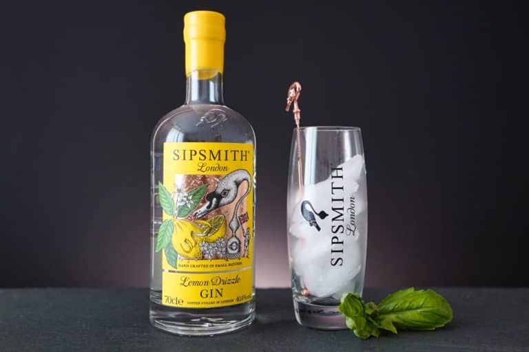 Neben einem hohen Glas mit Eis, einem Rührstäbchen und dem unverwechselbaren Sipsmith Lemon Drizzle Gin-Logo steht eine Flasche Sipsmith Lemon Drizzle Gin. Vor dem Glas liegt auf einer dunklen Unterlage ein Basilikumzweig.