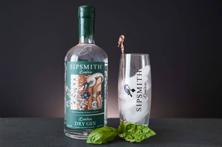 Neben einem Glas mit Eis und einem Rührstäbchen aus Kupfer, das mit „Sipsmith London“ beschriftet ist, steht eine Flasche Sipsmith Gin London Dry. Vorne liegen zwei grüne Basilikumblätter auf einer dunklen Fläche vor schwarzem Hintergrund.