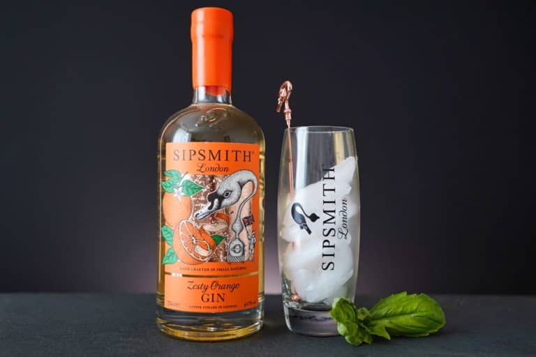 Neben einem hohen Glas mit Eis, einem Metallrührstäbchen und einem Sipsmith-Logo steht eine Flasche Sipsmith Zesty Orange Gin. Davor liegen frische Basilikumblätter – alles auf einer dunklen Oberfläche vor schwarzem Hintergrund –, was den perfekten Rahmen bildet, um den lebendigen Charakter des Sipsmith Zesty Orange Gin zur Geltung zu bringen.