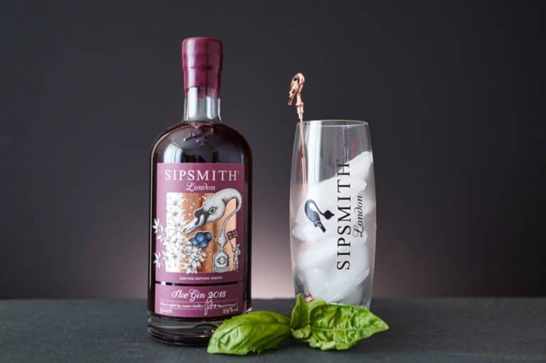 Neben einem mit Eis gefüllten Glas der Marke Sipsmith, in dem ein Kupferlöffel liegt, steht eine Flasche Sipsmith Sloe Gin. Vor dem Glas liegen frische Basilikumblätter auf einer dunklen Unterlage, wodurch die satten Farbtöne des Sipsmith Sloe Gin vor dem schlichten schwarzen Hintergrund besonders gut zur Geltung kommen.