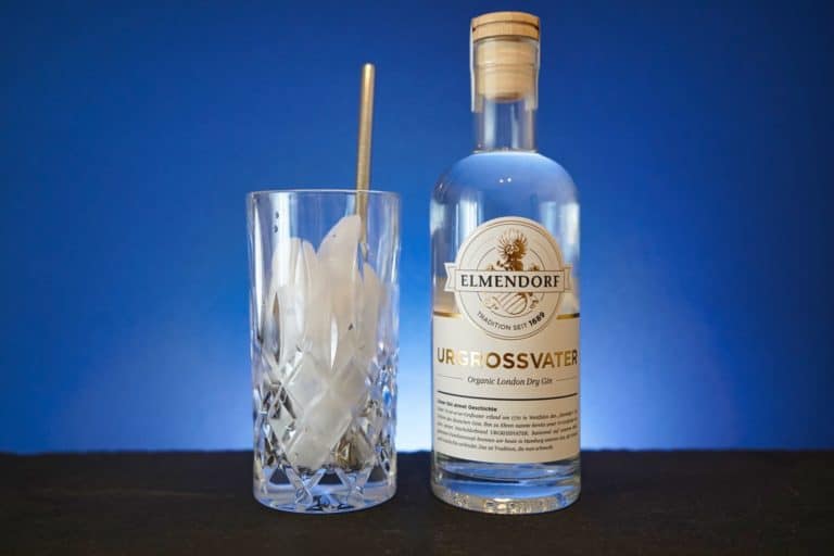 Vor einem blauen Hintergrund steht eine Flasche „Elmendorf Ur-Großvater Gin Bio“ neben einem mit Eis gefüllten Kristallglas und einem Metallstrohhalm.