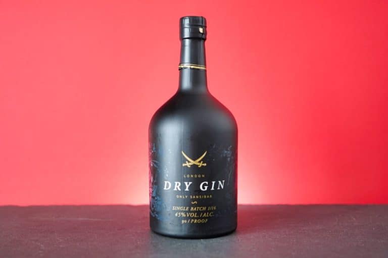 Eine mattschwarze Flasche Sansibar Gin, ein klassischer London Dry Gin mit goldenen Akzenten, steht auf einer dunklen Fläche vor einem leuchtend roten Hintergrund.
