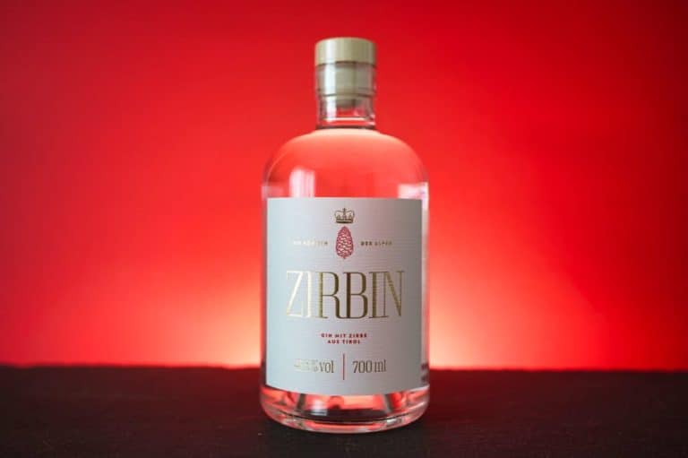 Eine durchsichtige Flasche Zirbin Gin mit einem weiß-goldenen Etikett steht auf einer schwarzen Fläche vor einem leuchtend roten Hintergrund.