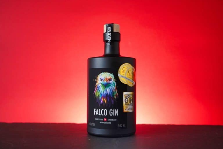 Eine elegante schwarze Flasche Falco Gin mit einer farbenfrohen Falkenillustration und Auszeichnungsaufklebern steht auf einer schwarzen Fläche vor einem auffälligen roten Farbverlaufshintergrund.