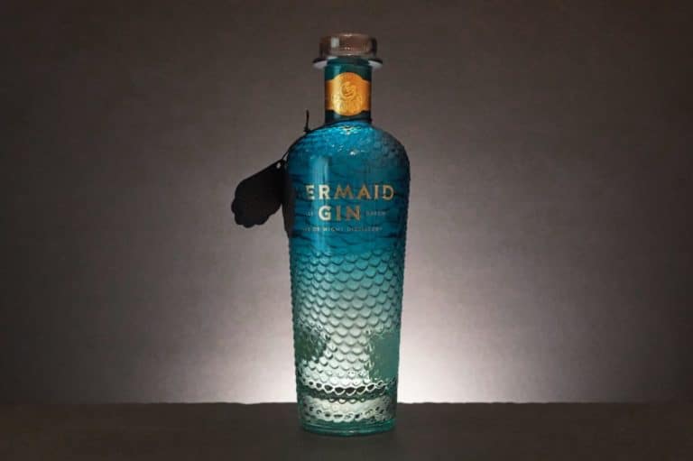 Eine auffällige blaue Flasche „Mermaid Gin“ mit einer strukturierten, schuppenartigen Oberfläche steht aufrecht vor einem dunklen Hintergrund und wird von hinten beleuchtet.