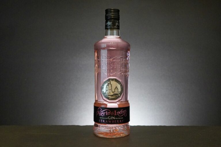 Eine Flasche „Puerto de Indias Strawberry Gin“ mit ihrer charakteristischen rosa Flüssigkeit, dem schwarzen Verschluss und dem schwarzen Etikett steht elegant auf einer dunklen Fläche vor einem grauen Hintergrund mit Farbverlauf.