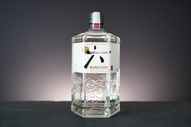 Auf einer schwarzen Fläche vor einem dunklen Hintergrund steht eine Flasche Roku Gin mit weißem Etikett und japanischer Aufschrift, sechseckiger Form und floralen Gravuren.