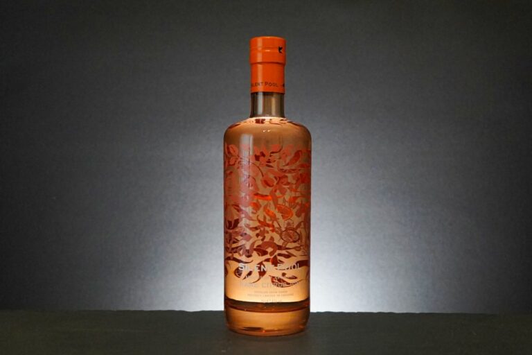 Eine Flasche mit klarem Spirituosengetränk, die als „Silent Pool Rare Citrus Gin“ bezeichnet wird, verfügt über einen orangefarbenen Verschluss und dekorative orangefarbene Blumenmuster auf dem Glas, die sich vor einem dunklen Hintergrund abheben und von hinten leicht beleuchtet werden.
