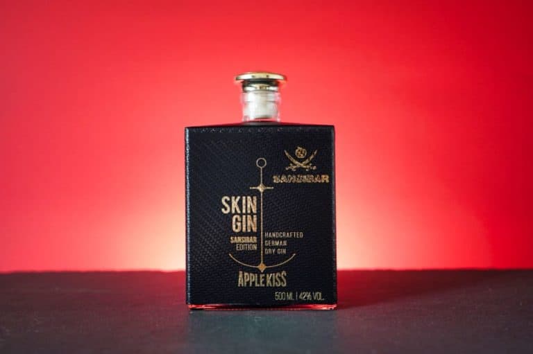 Eine schwarze Flasche „Skin Gin Apple Kiss“ mit goldener Beschriftung hebt sich von einem roten Hintergrund mit Farbverlauf ab und strahlt jene kühne Eleganz aus, die an „Gin Sansibar“ erinnert.